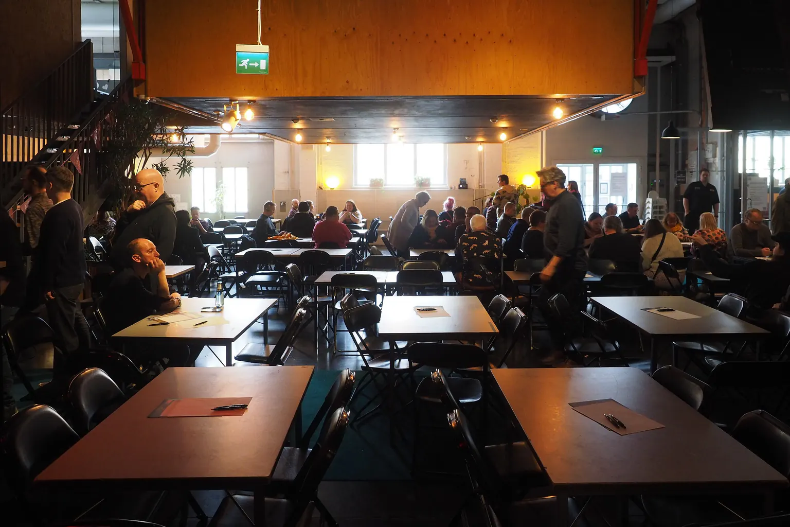 A large quiz room as the first teams arrive: answer sheets and pens laid out on empty tables, a few people seated at the back.