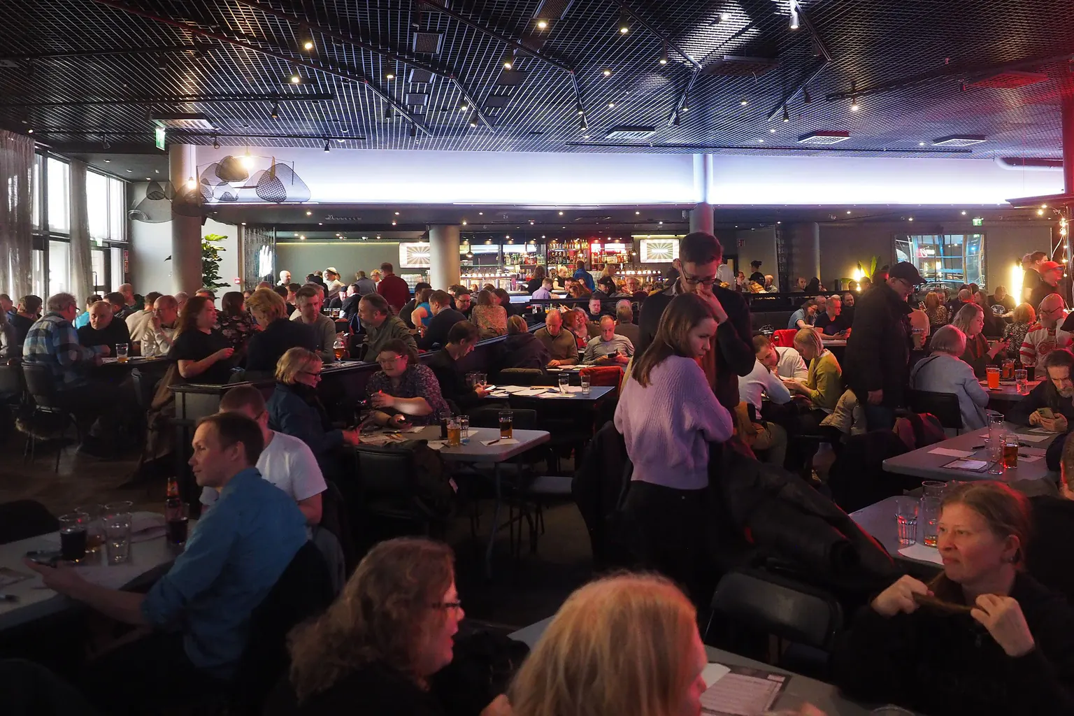 A different large room during a quiz, with teams at every table and a few people standing between them.
