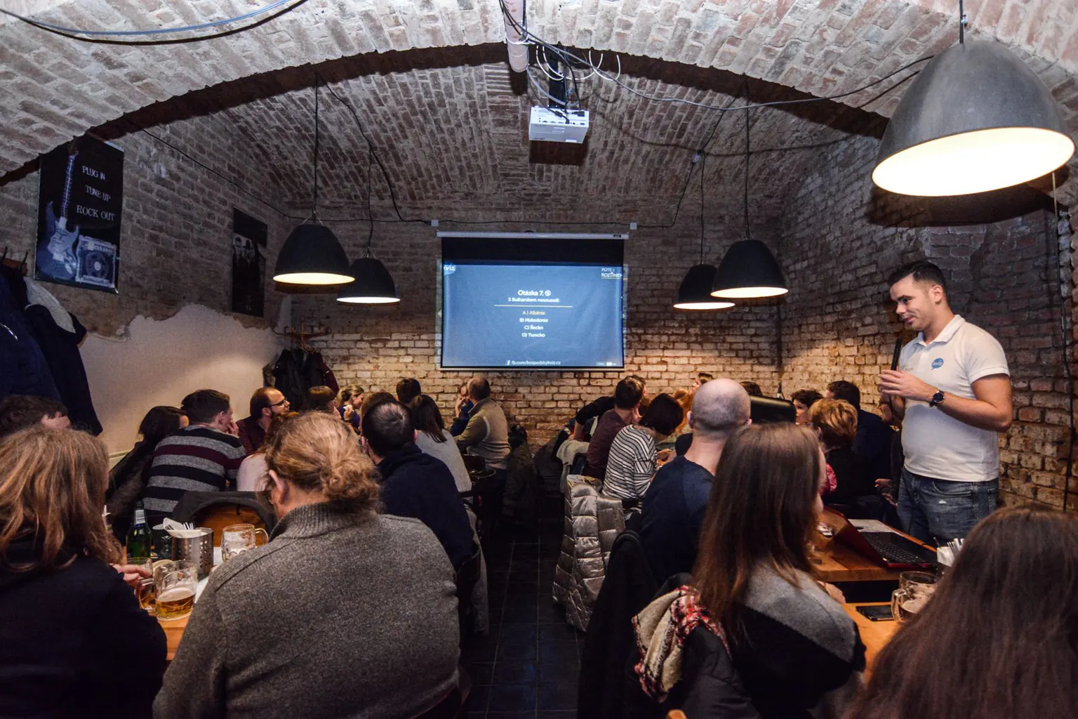 A quiz host holding a microphone talks to teams at long tables in a brick-vaulted cellar bar, with a question on the projector screen behind him.