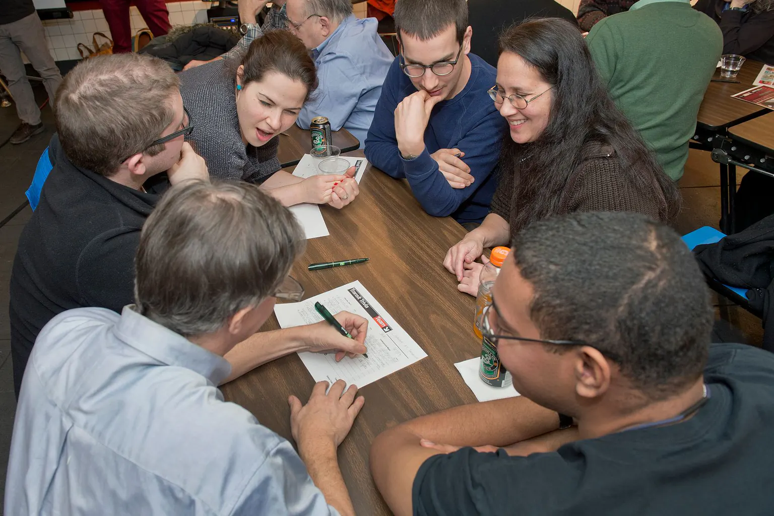 A team of six leans in around a table: one writes on the answer sheet while another makes her case.