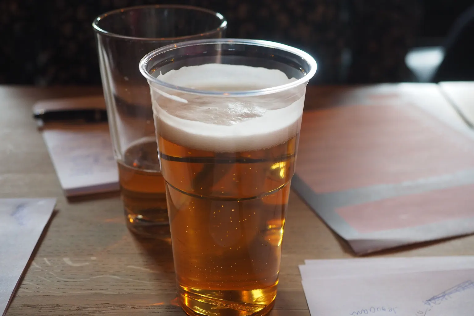 A plastic cup of beer on a wooden table between quiz answer sheets and a pen.