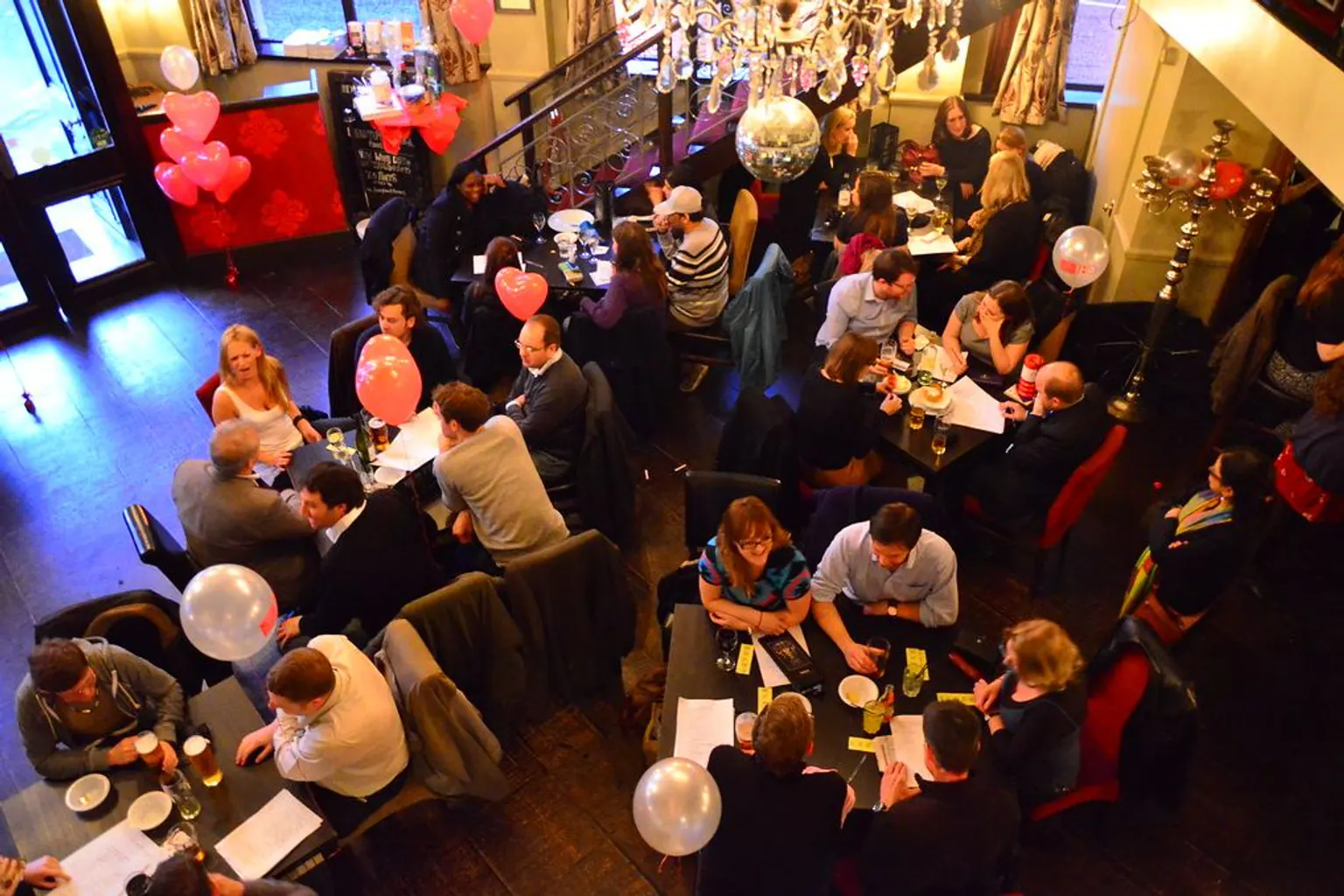 Seen from above, teams sit at tables across a pub during a quiz, with balloons tied to the chairs.