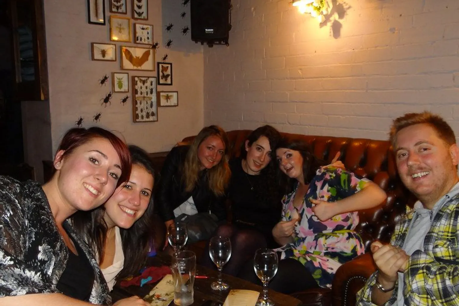 A quiz team squeezes onto a leather sofa to celebrate, smiling at the camera, with wine glasses on the table.